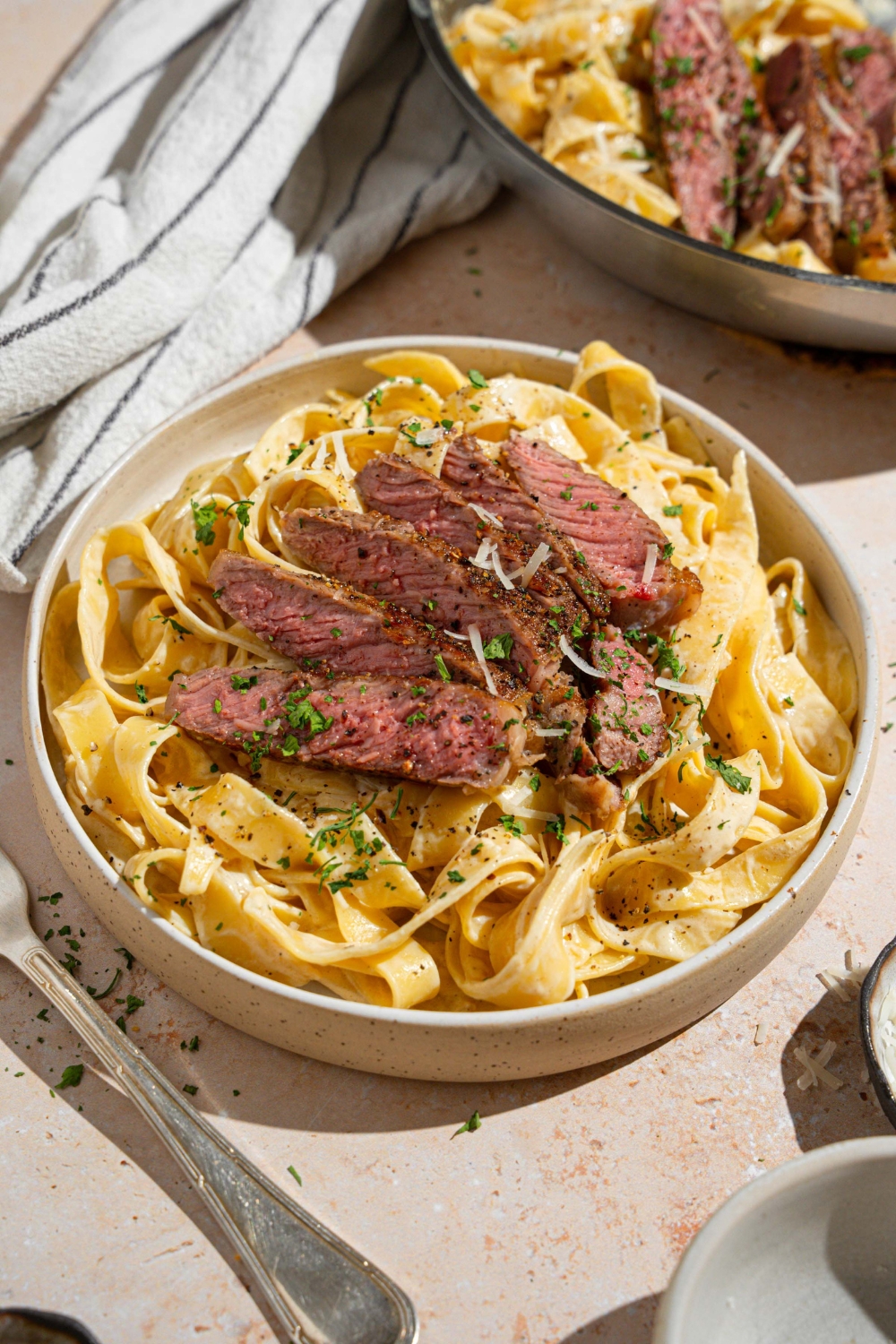 Fettuccine alfredo topped with sliced steak garnished with fresh parsley and grated cheese in a white bowl. The bowl is on a tan counter with a fork and white striped napkin.