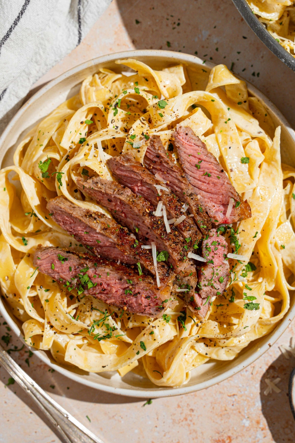 Fettuccine alfredo topped with sliced steak garnished with fresh parsley and grated cheese in a white bowl. The bowl is on a tan counter with a fork and white striped napkin.