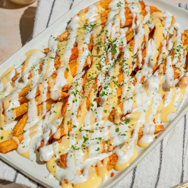 A white platter with copycat Wingstop Voodoo fries drizzled with cheese and ranch dressing. The plate is garnished with chopped parsley and served over a white striped napkin.