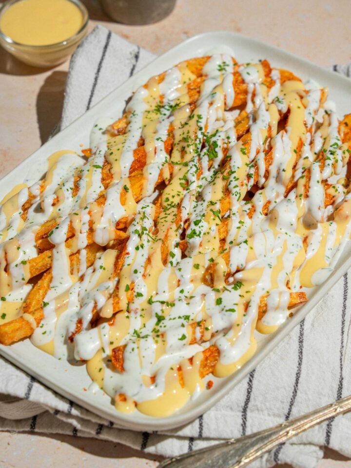 A white platter with copycat Wingstop Voodoo fries drizzled with cheese and ranch dressing. The plate is garnished with chopped parsley and served over a white striped napkin.