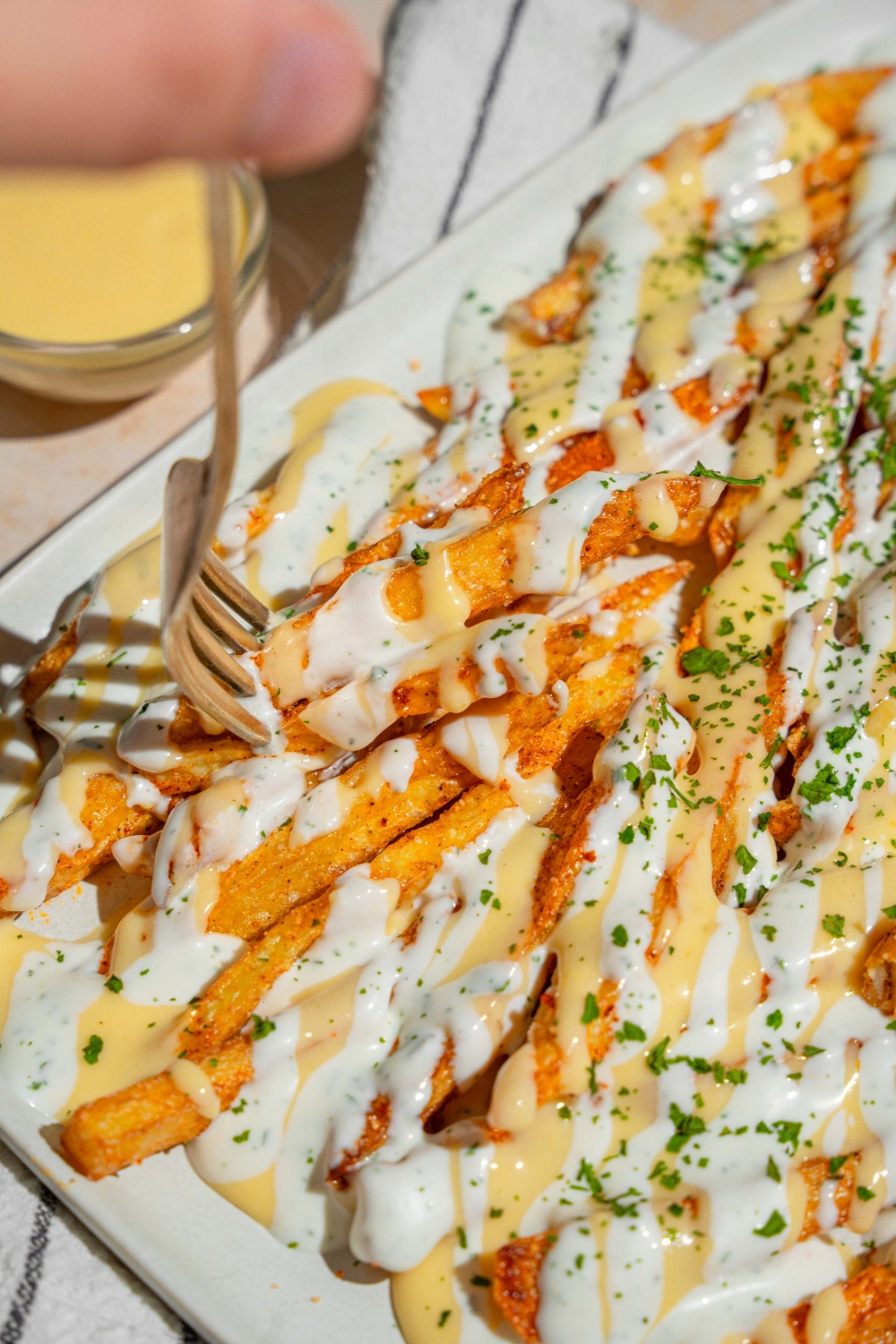 A white platter with copycat Wingstop Voodoo fries drizzled with cheese and ranch dressing. The plate is garnished with chopped parsley and served over a white striped napkin. A fork is taking a bite from the fries.