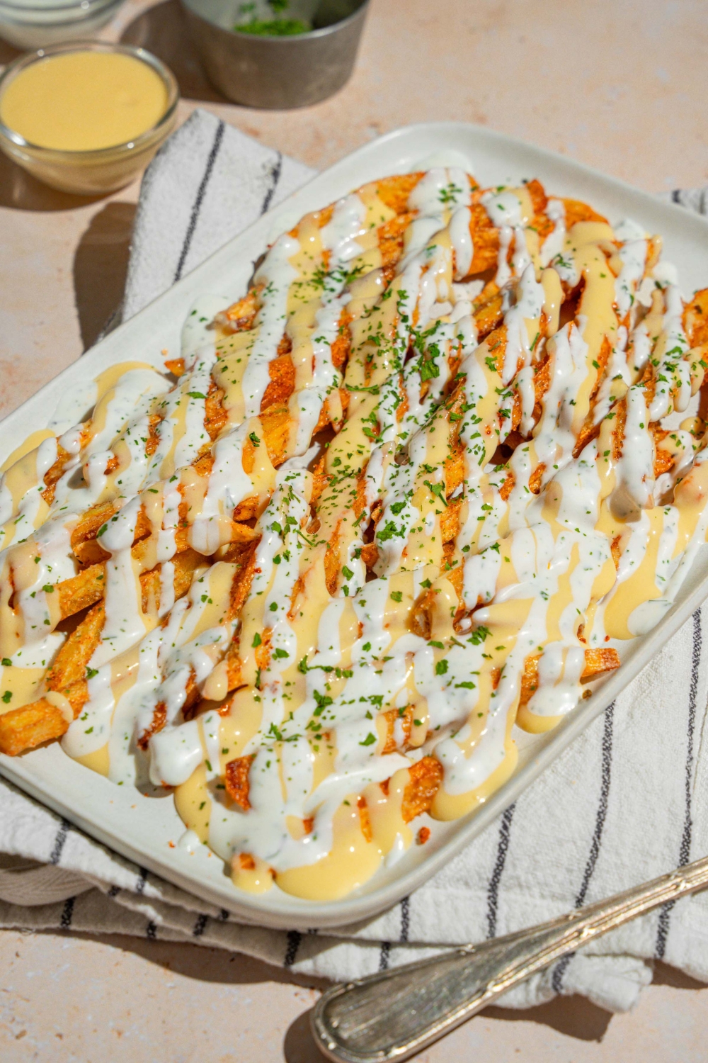 A white platter with copycat Wingstop Voodoo fries drizzled with cheese and ranch dressing. The plate is garnished with chopped parsley and served over a white striped napkin.
