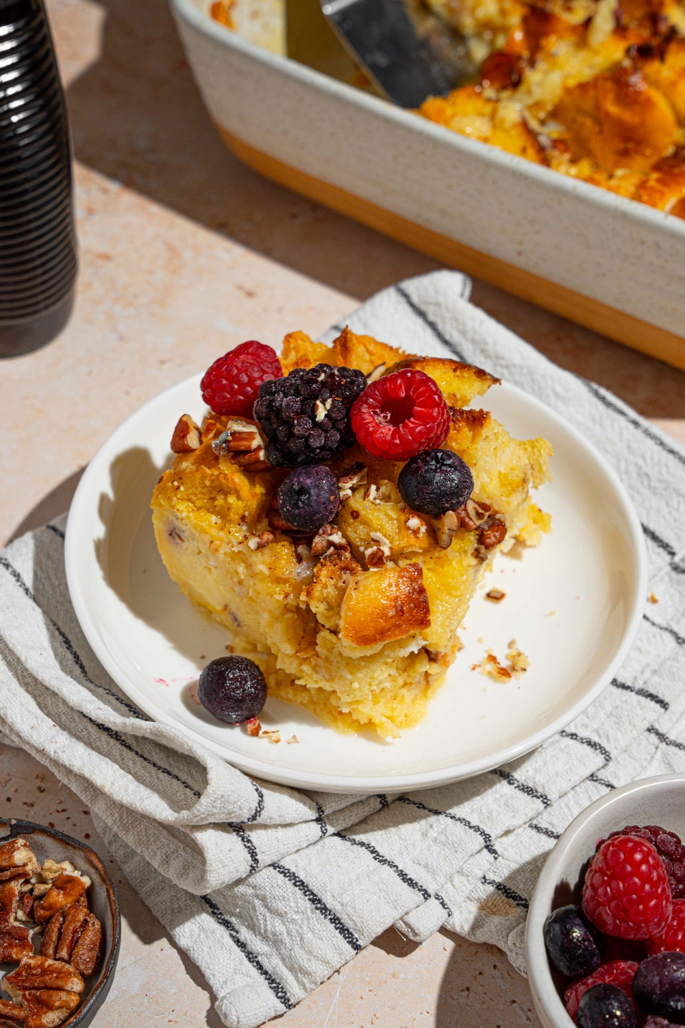 A white plate with a slice of challah french toast topped with mixed berries. The plate is on a tan counter with a white striped napkin with a bowl of berries and plate of pecans.