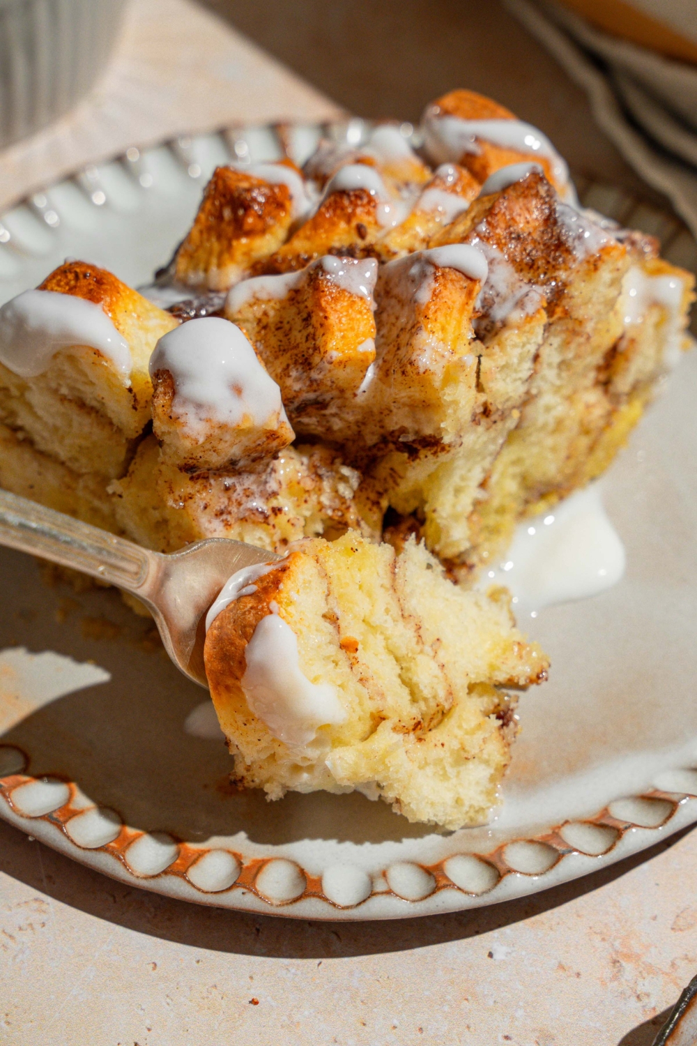 A ceramic plate with a slice of cinnamon roll french toast drizzled with icing. There is a fork on the plate with a bite of french toast. The plate is on a tan counter.