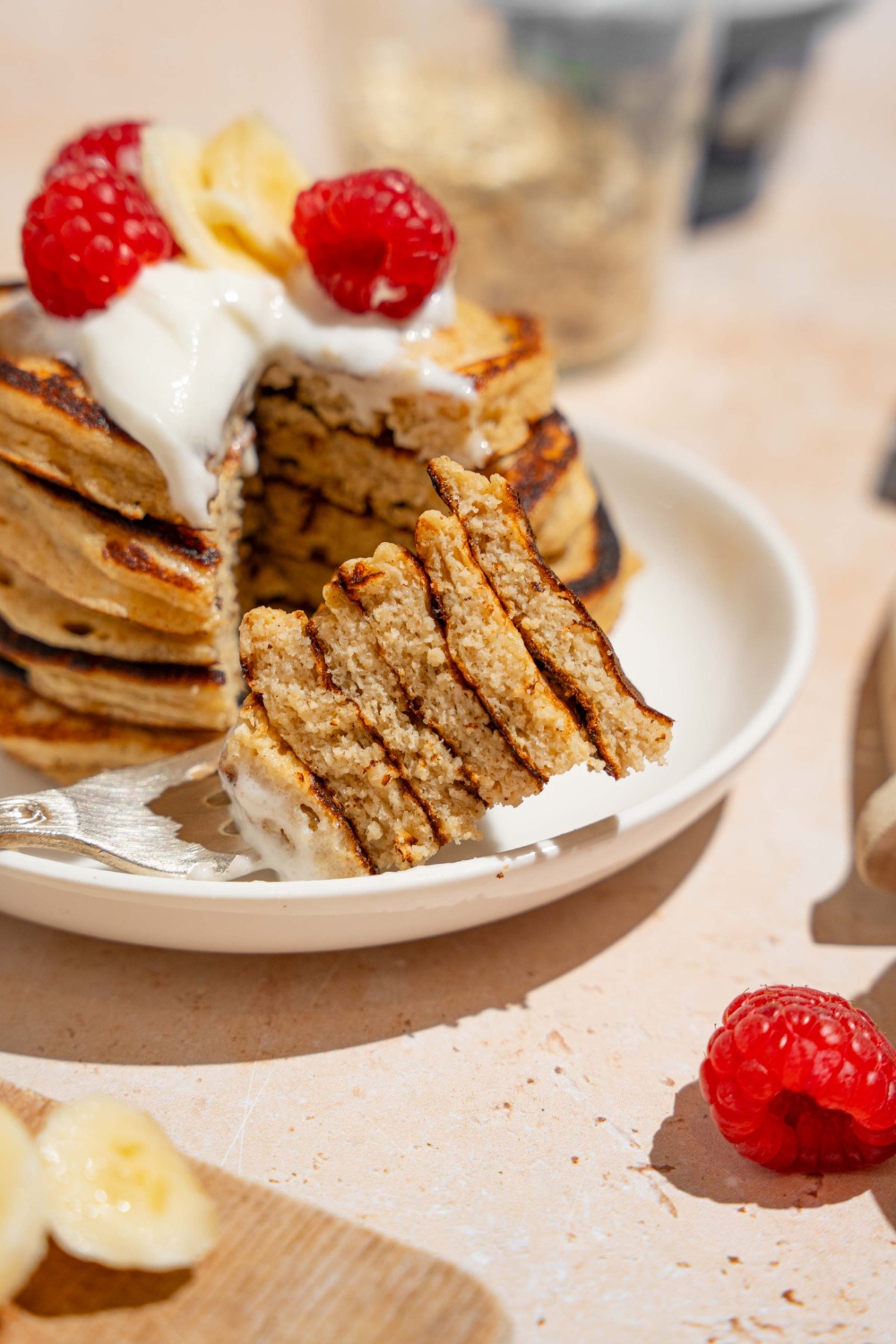 A white plate with a stack of cottage cheese banana pancakes topped with whipped cream, sliced banana, and raspberries. A fork has taken a bite from the stack. The plate is on a tan counter with bananas and a bowl of raspberries.