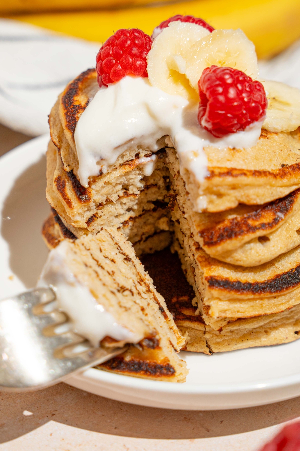 A white plate with a stack of cottage cheese banana pancakes topped with whipped cream, sliced banana, and raspberries. A fork has taken a bite from the stack. The plate is on a tan counter.