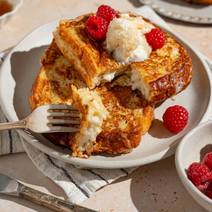 A white plate with stuffed french toast topped with whipped cream cheese and raspberries drizzled with syrup. There is a fork taking a bite of french toast. The plate is on a tan counter with a white striped napkin, bowl of syrup, and bowl of raspberries.