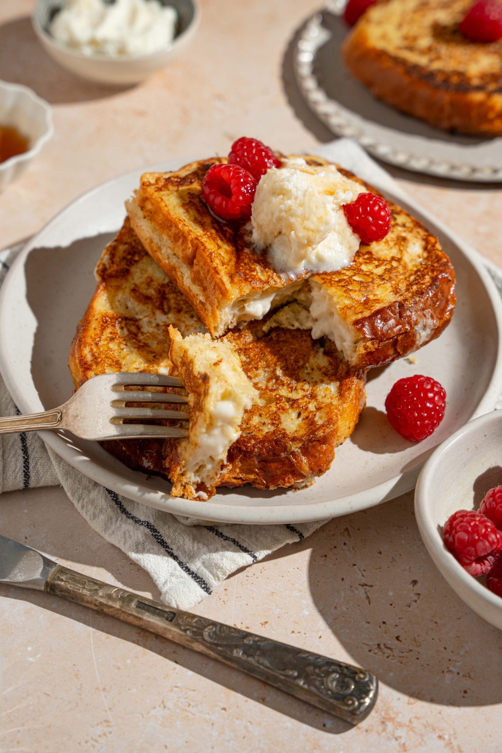 A white plate with stuffed french toast topped with whipped cream cheese and raspberries drizzled with syrup. There is a fork taking a bite of french toast. The plate is on a tan counter with a white striped napkin, bowl of syrup, and bowl of raspberries.