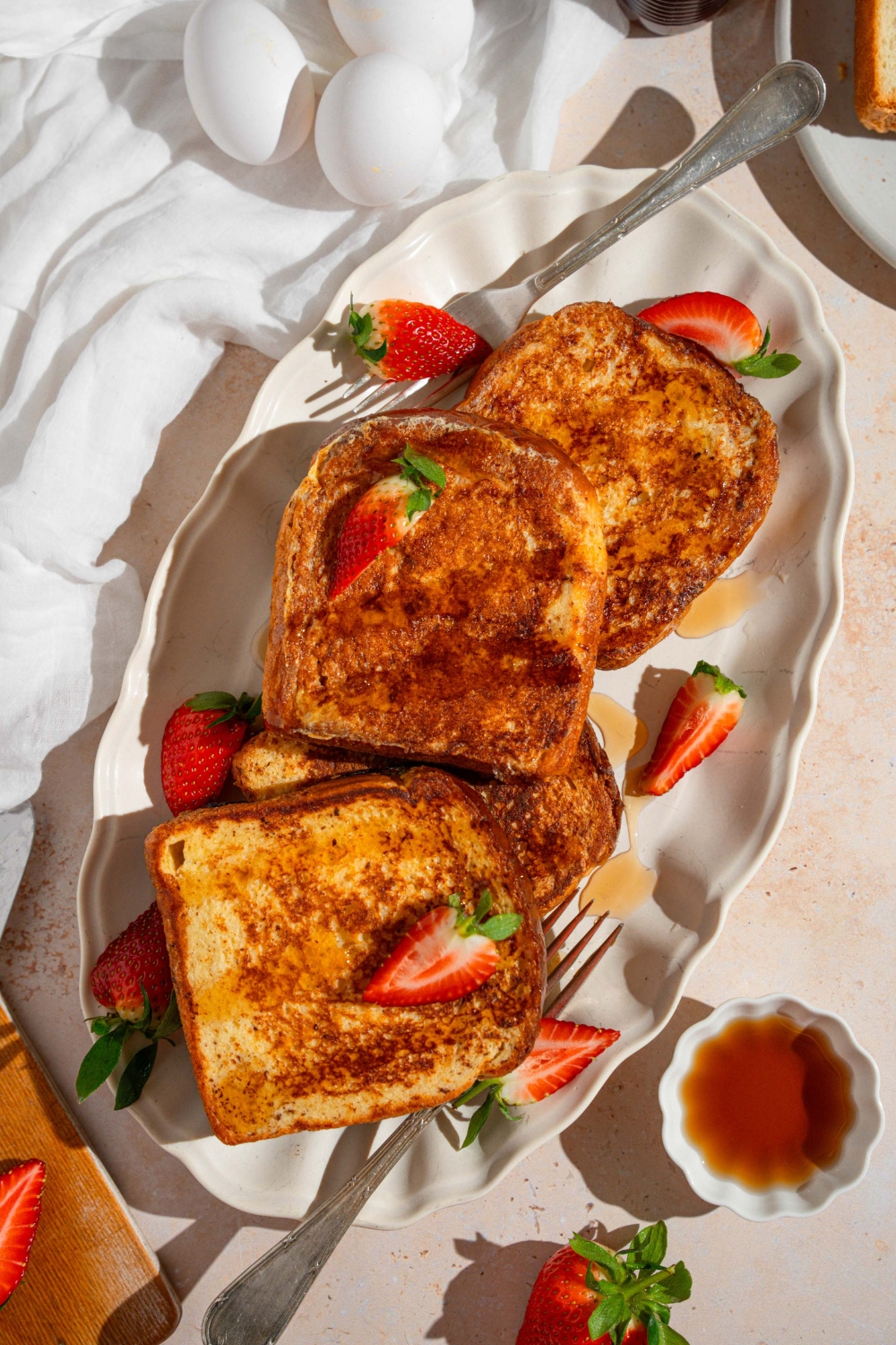 A white platter with slices of French toast drizzled with maple syrup and garnished with sliced strawberries. There is a fork on the plate. The plate is on a tan counter with a white cloth napkin, eggs, and small ramekin of syrup.