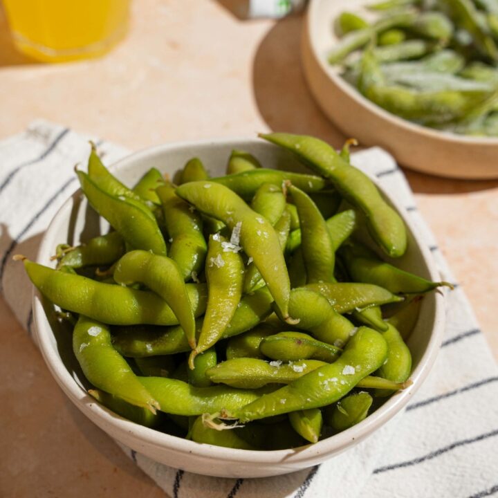 A bowl of cooked edamame seasoned with salt. The bowl is on a white striped napkin on a tan counter with a plate of frozen edamame.