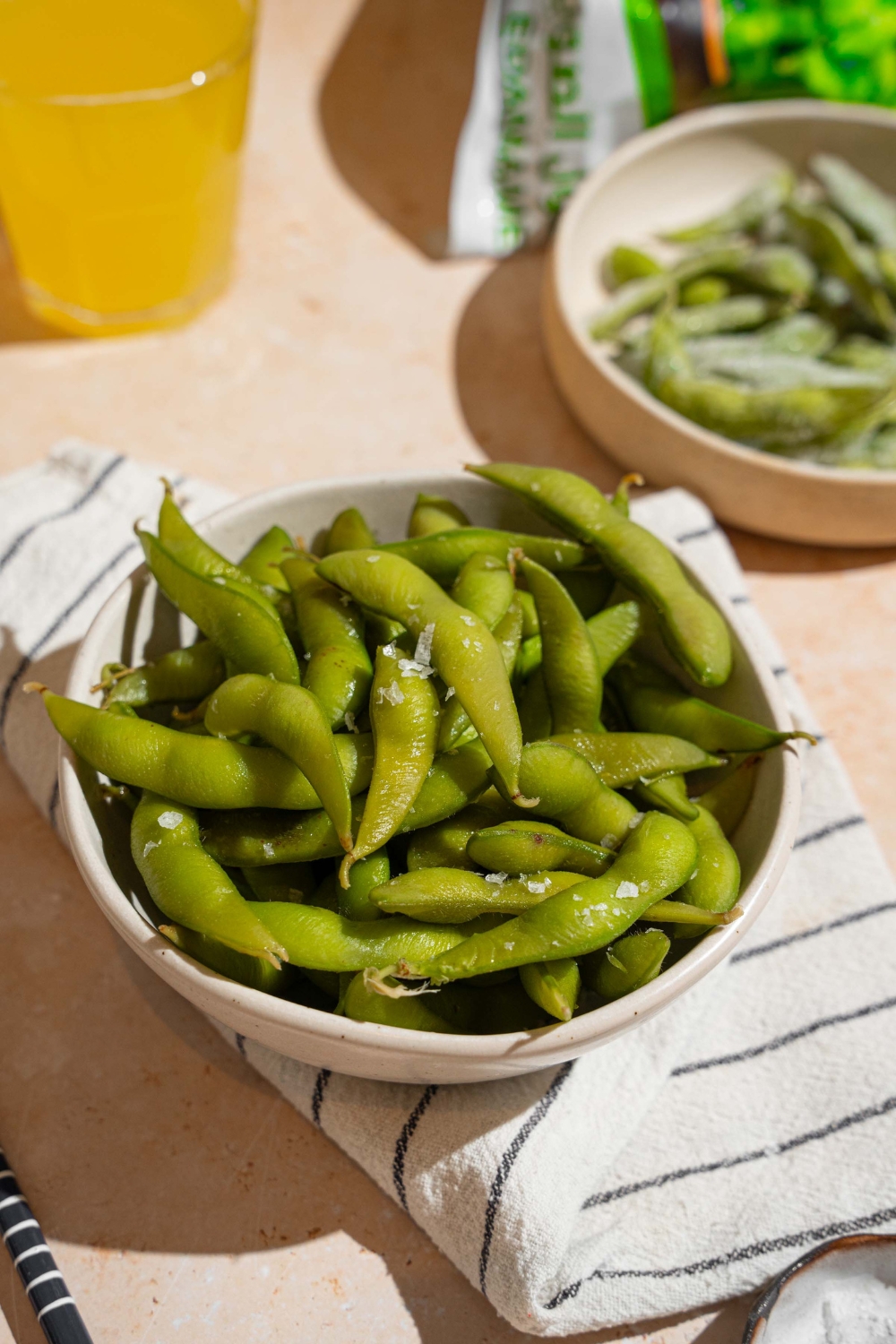 A bowl of cooked edamame seasoned with salt. The bowl is on a white striped napkin on a tan counter with a plate of frozen edamame.