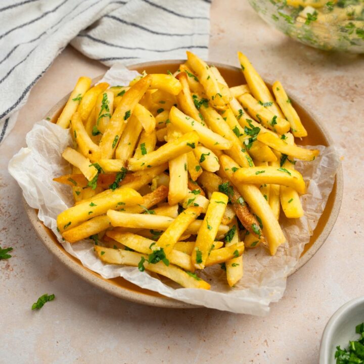 Garlic fries garnished with fresh parsley served on a plate lined with parchment paper. The plate is on a tan counter with a white striped napkin and glass bowl with fries.