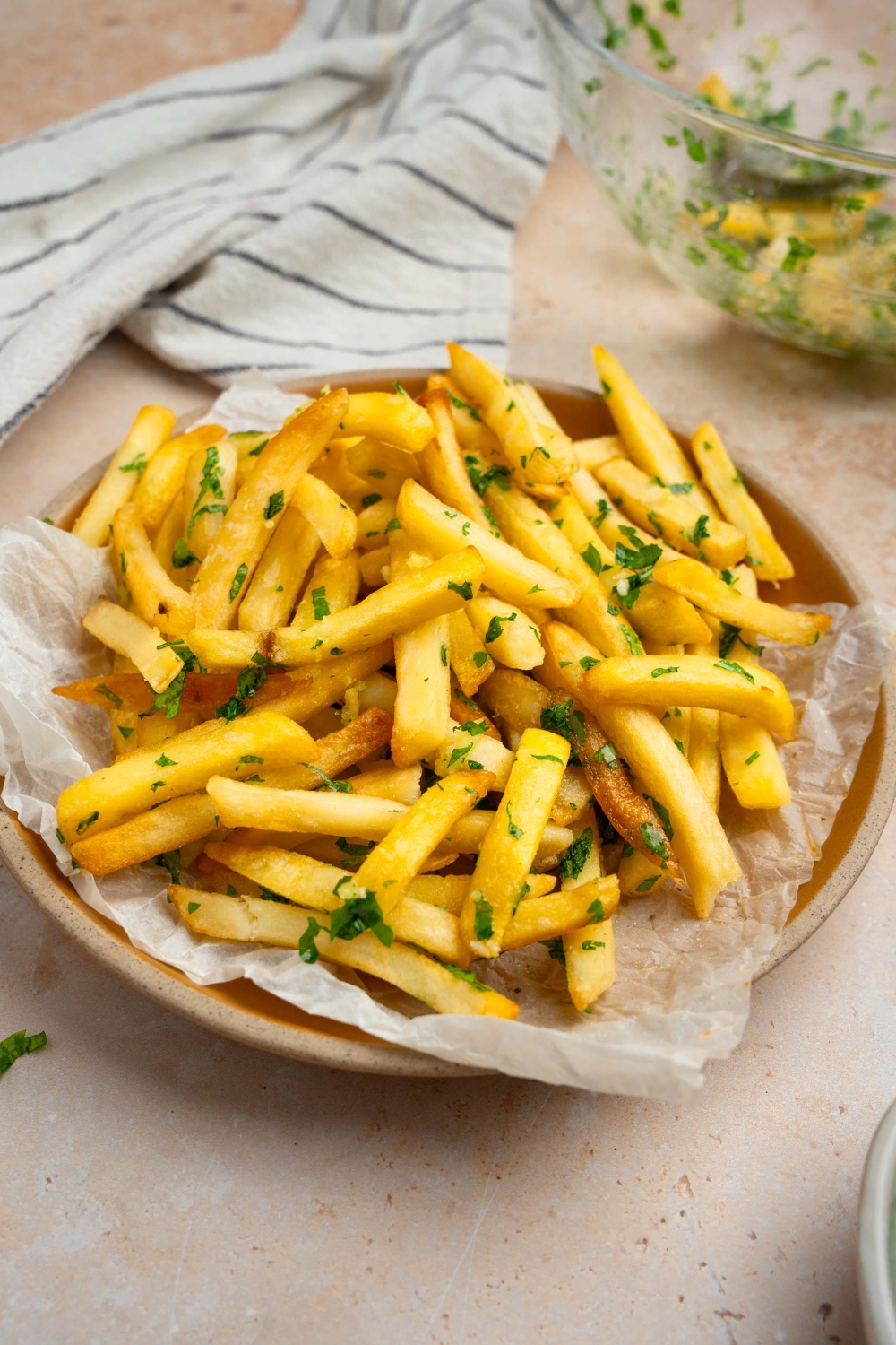 Garlic fries garnished with fresh parsley served on a plate lined with parchment paper. The plate is on a tan counter with a white striped napkin.