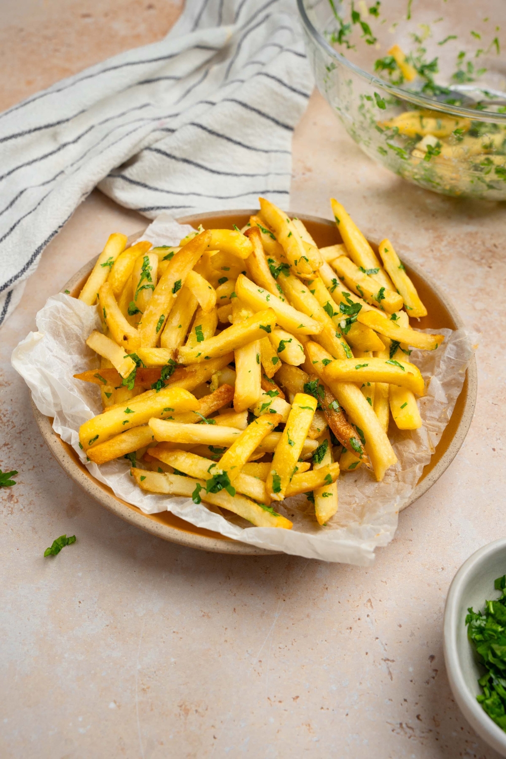 Garlic fries garnished with fresh parsley served on a plate lined with parchment paper. The plate is on a tan counter with a white striped napkin and glass bowl with fries.