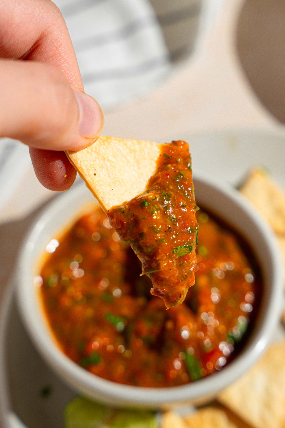 A close up of a tortilla chip dipped in homemade Chipotle red chimichurri sauce. There is a plate of chips with a bowl of sauce blurred in the background.