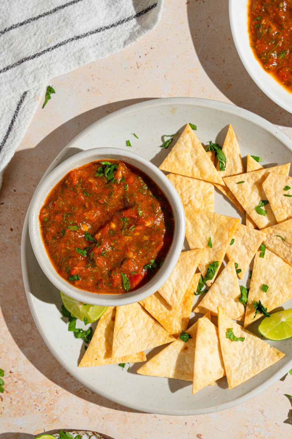 A bowl of Chipotle red chimichurri garnished with fresh parsley on a plate with tortilla chips. The plate is served with a lime wedge on a tan counter with a white striped napkin and bowl of chimichurri.