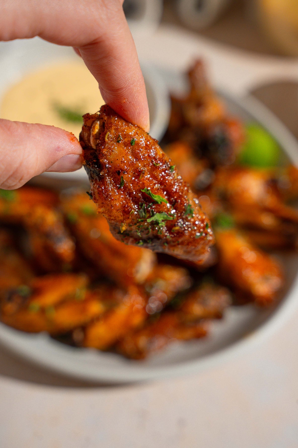 A close up of a hand holding a copycat Wingstop Louisiana rub wing garnished with fresh parsley. There is a plate of wings with wing sauce blurred in the background.