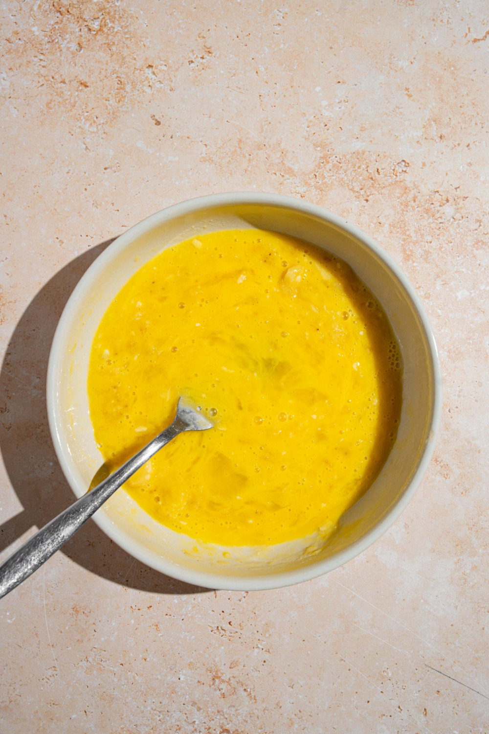 A large white bowl with a fork mixing mashed bananas with whisked eggs. The bowl is on a tan counter.