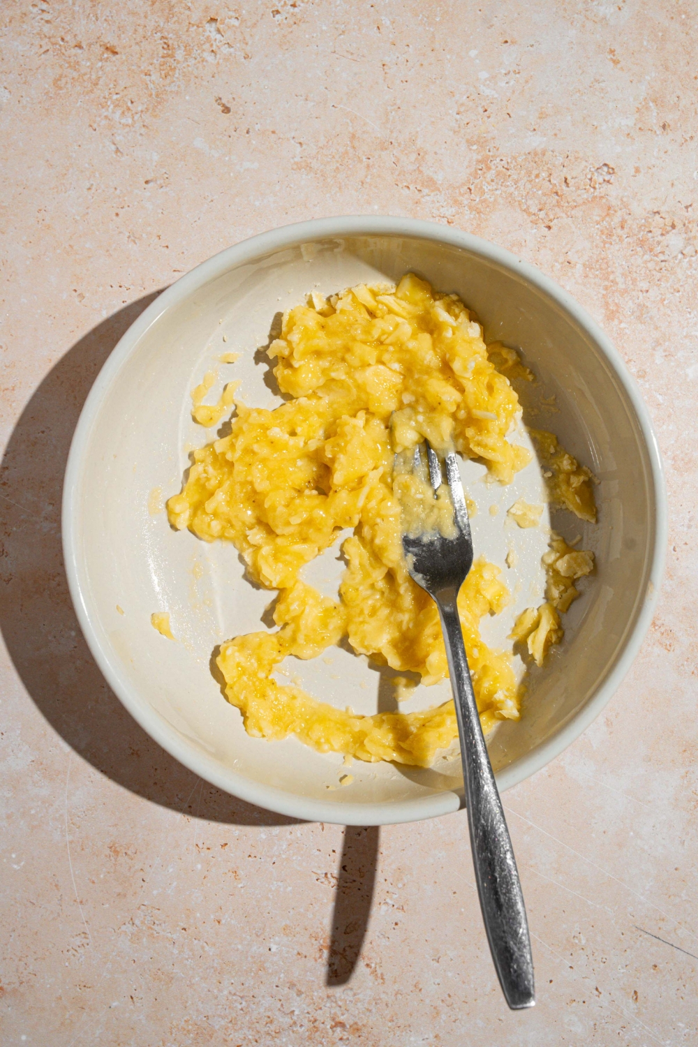 A white bowl with a fork mashing bananas. The bowl is on a tan counter.
