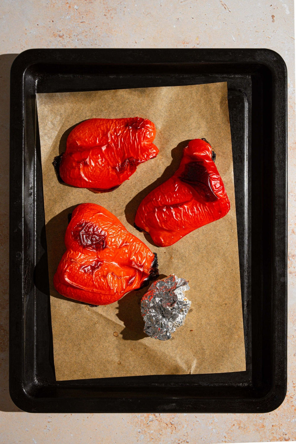 A baking sheet lined with parchment paper with roasted halved red peppers and garlic wrapped in foil. The baking sheet is on a tan counter.