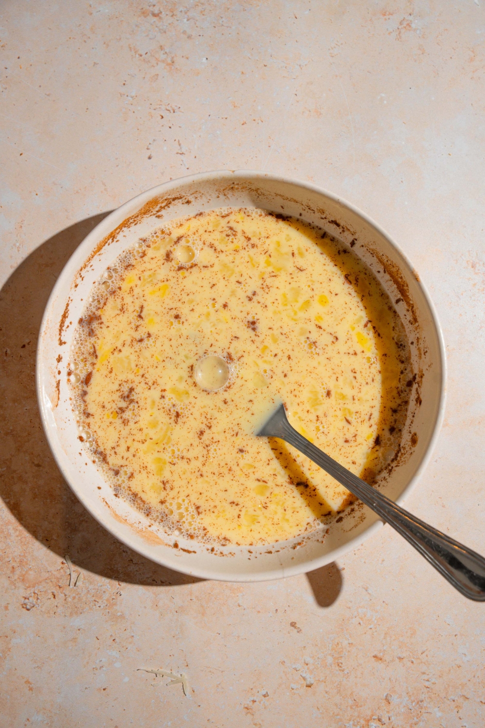 A white bowl with fork combining ingredients to make french toast including milk, eggs, cinnamon, vanilla, and sugar. The bowl is on a tan counter.