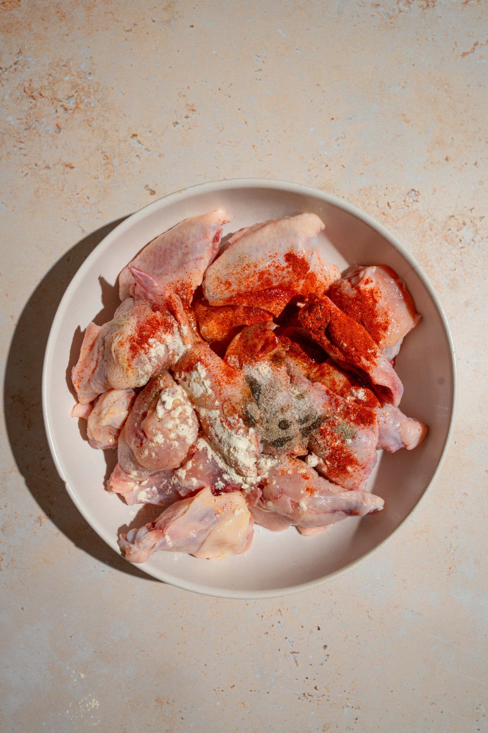 A white bowl with uncooked wings topped with seasonings to make a Wingstop Louisiana rub. The bowl is on a tan counter.
