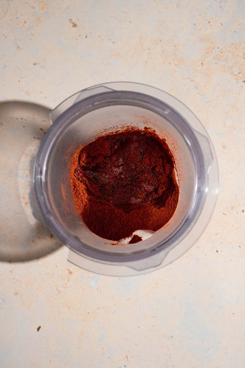 A blender jar with ingredients to make adobo sauce including chile powder, salt, oregano, cinnamon, and cumin. The jar is on a tan counter.