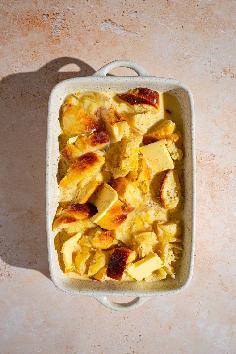 A baking dish with pieces of challah bread topped with butter and soaking in a milk mixture. The dish is on tan counter.