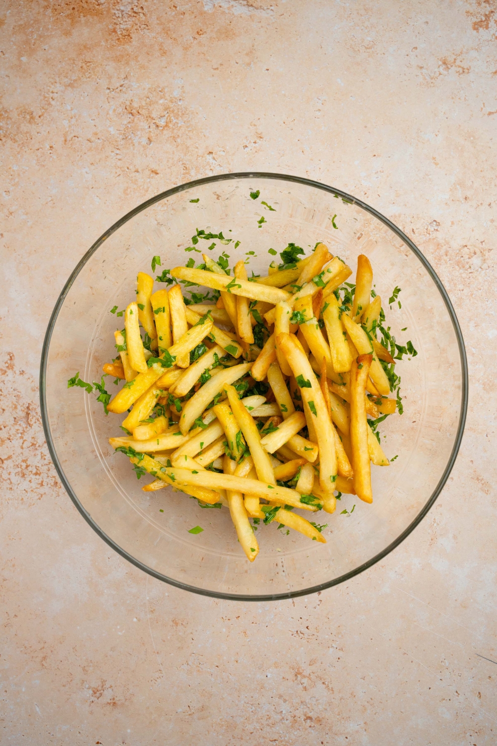 A glass bowl with french fries tossed in seasonings. The bowl is on a tan counter.