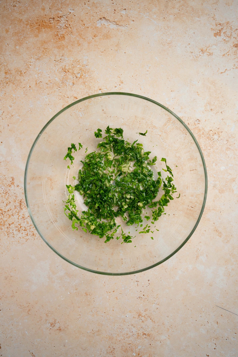 A glass bowl with fresh parsley mixed with garlic and oil. The bowl is on a tan counter.