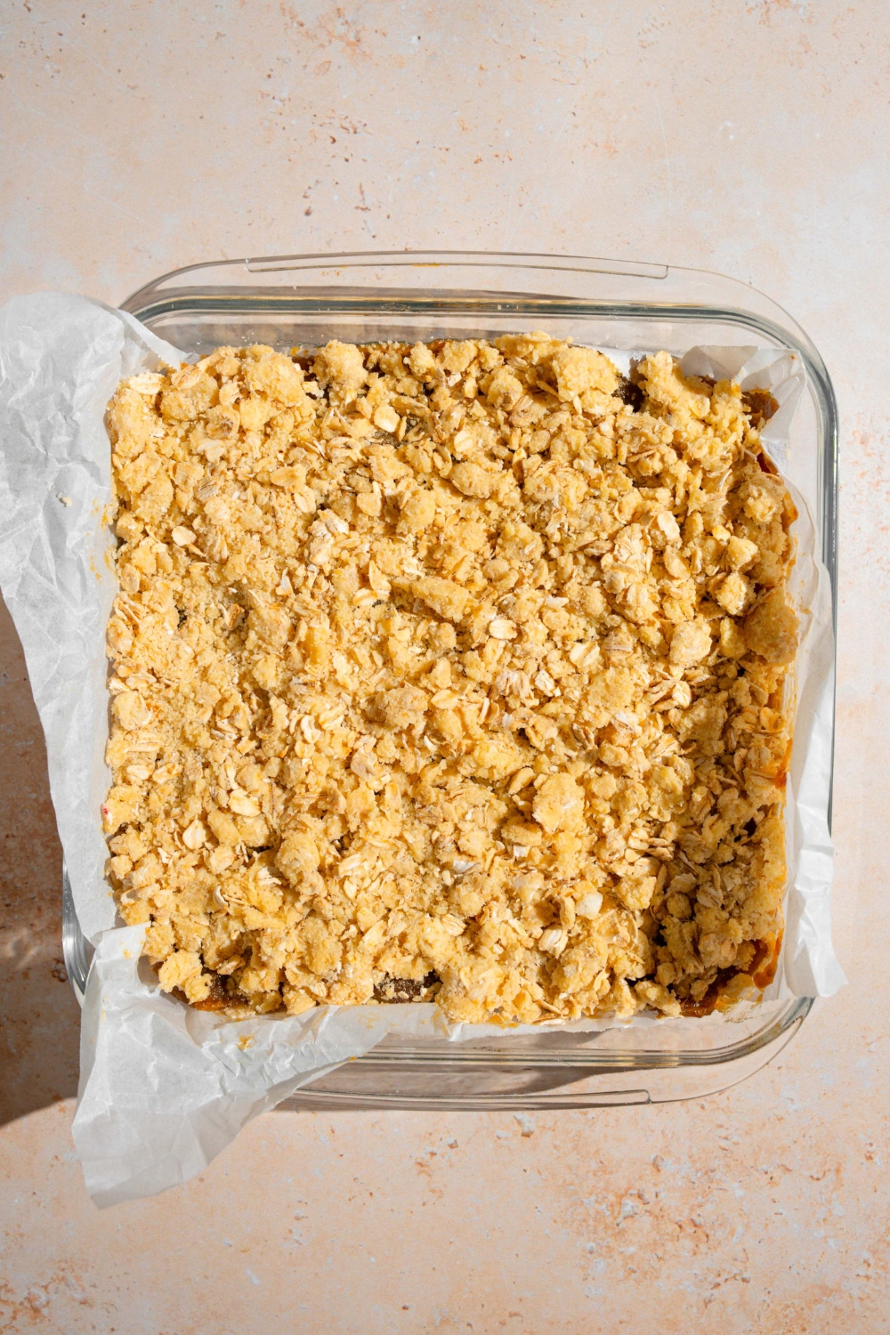 A glass baking dish lined with parchment paper with uncooked old fashioned date bars. The baking dish is on a tan counter.