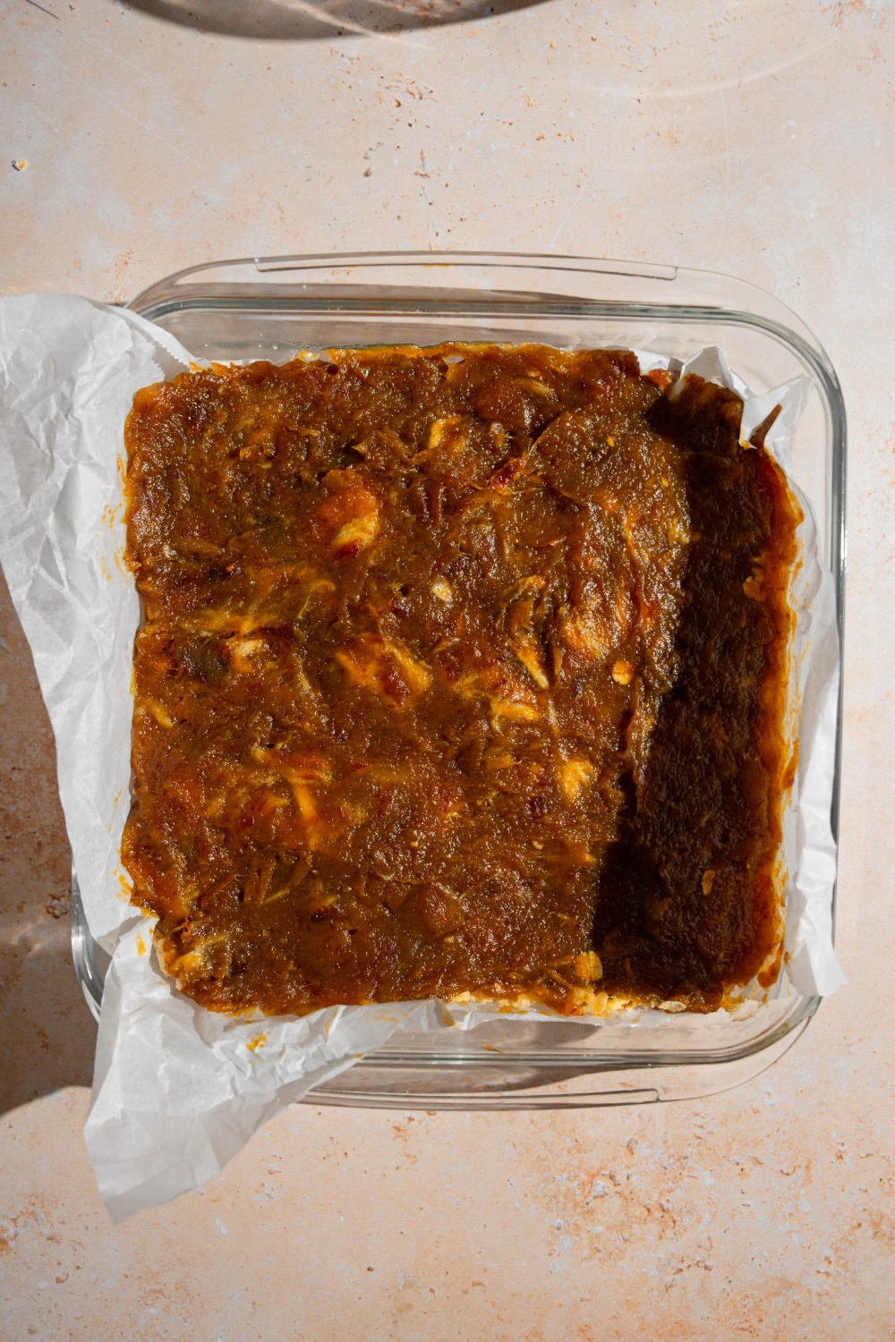A glass baking dish lined with parchment paper with uncooked crust topped with date filling. The baking dish is on a tan counter.