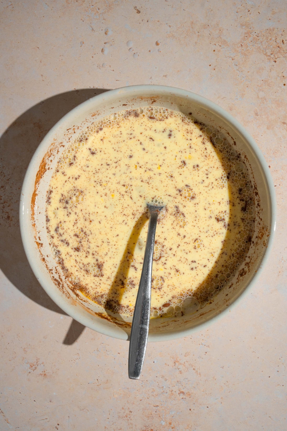 A bowl with a spoon mixing french toast egg mixture including eggs, milk, and cinnamon. The bowl is on a tan counter.