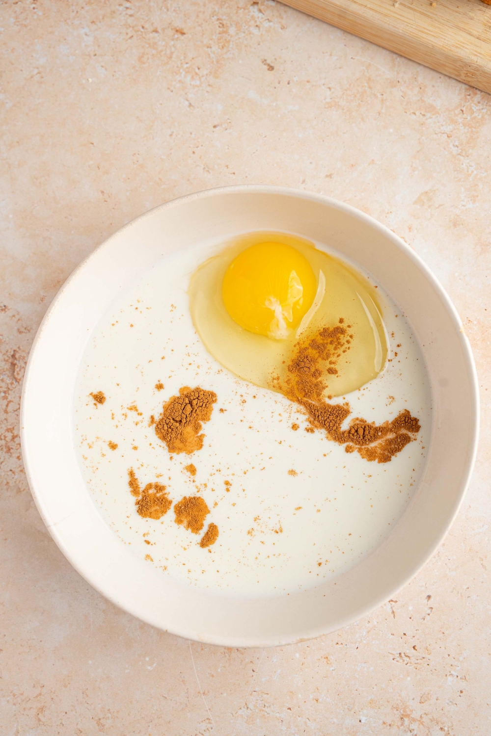 A white bowl with milk, eggs, cinnamon, and sugar. The bowl is on a tan counter.