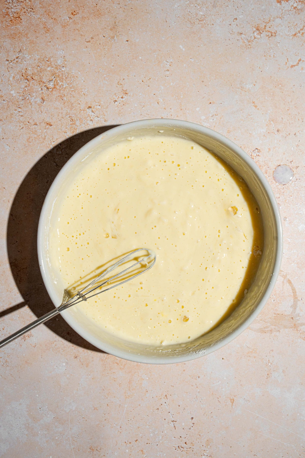 A white bowl with a whisk mixing sweet cream pancake batter. The bowl is on a tan counter.