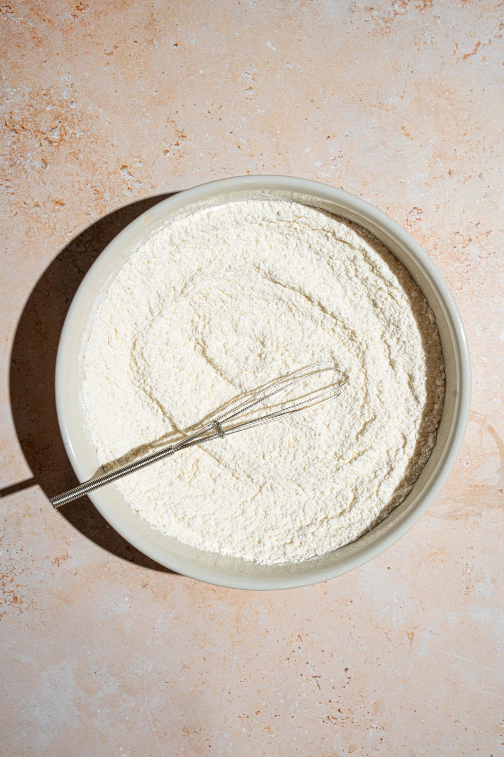 A white bowl with a whisk mixing dry ingredients to make sweet cream pancake batter. The bowl is on a tan counter.