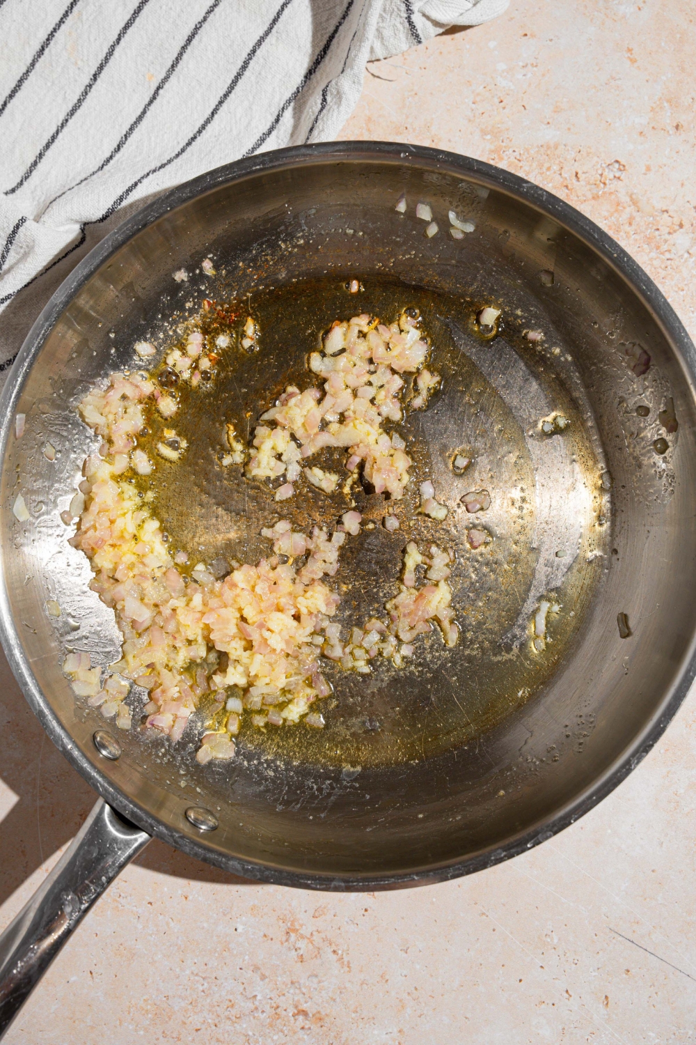 A skillet with garlic and shallots cooking in butter. The skillet is on a tan counter with a white striped napkin.