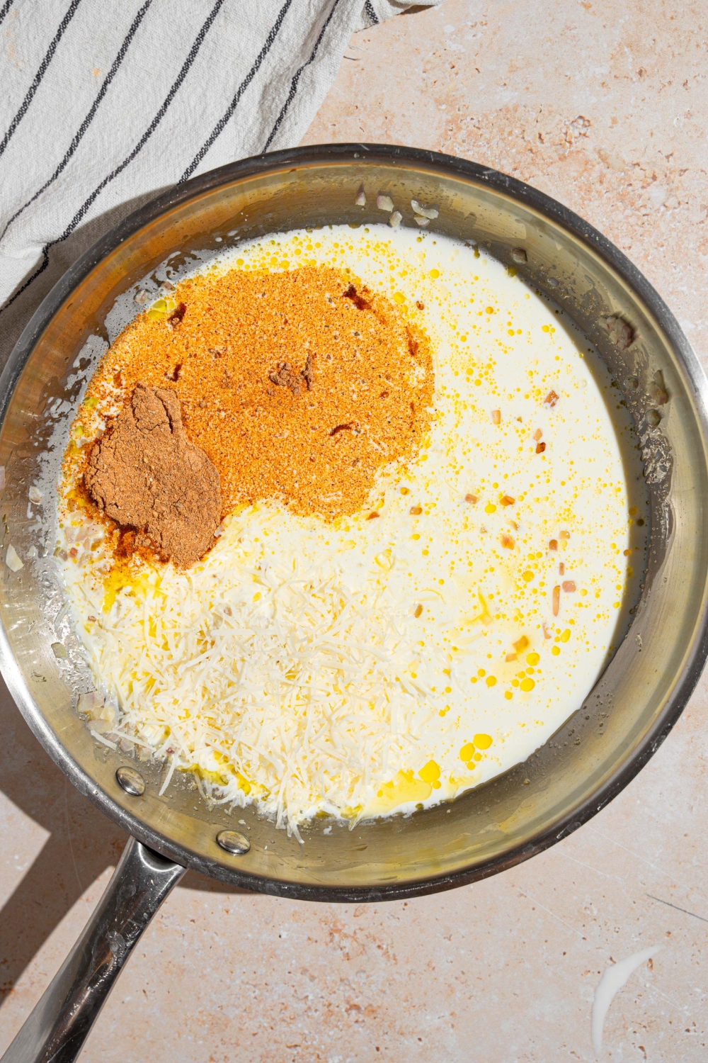 A skillet with ingredients to make cajun alfredo sauce including cajun seasoning, cream, and grated cheese. The skillet is on a tan counter with a white striped napkin.