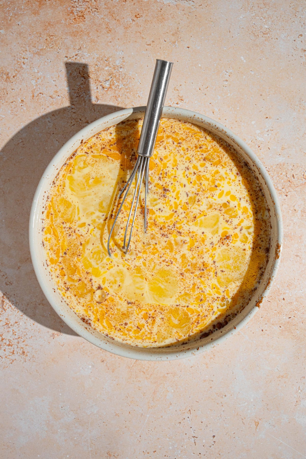 A white bowl with a spoon mixing a milk, egg, and cinnamon mixture. The bowl is on a tan counter.