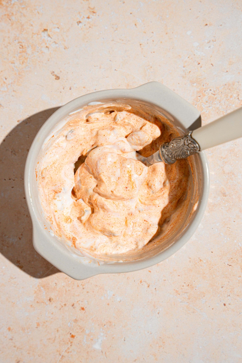 A ceramic ramekin with a spoon mixing a creamy jalapeno sauce. The bowl is on a tan counter.