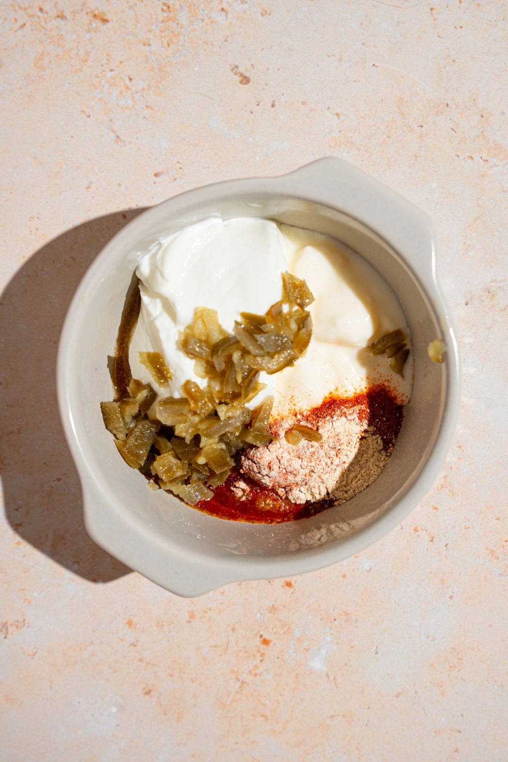 A ceramic ramekin with ingredients to make creamy jalapeno sauce including mayo, sour cream, jalapenos, paprika, cumin, and other seasonings.