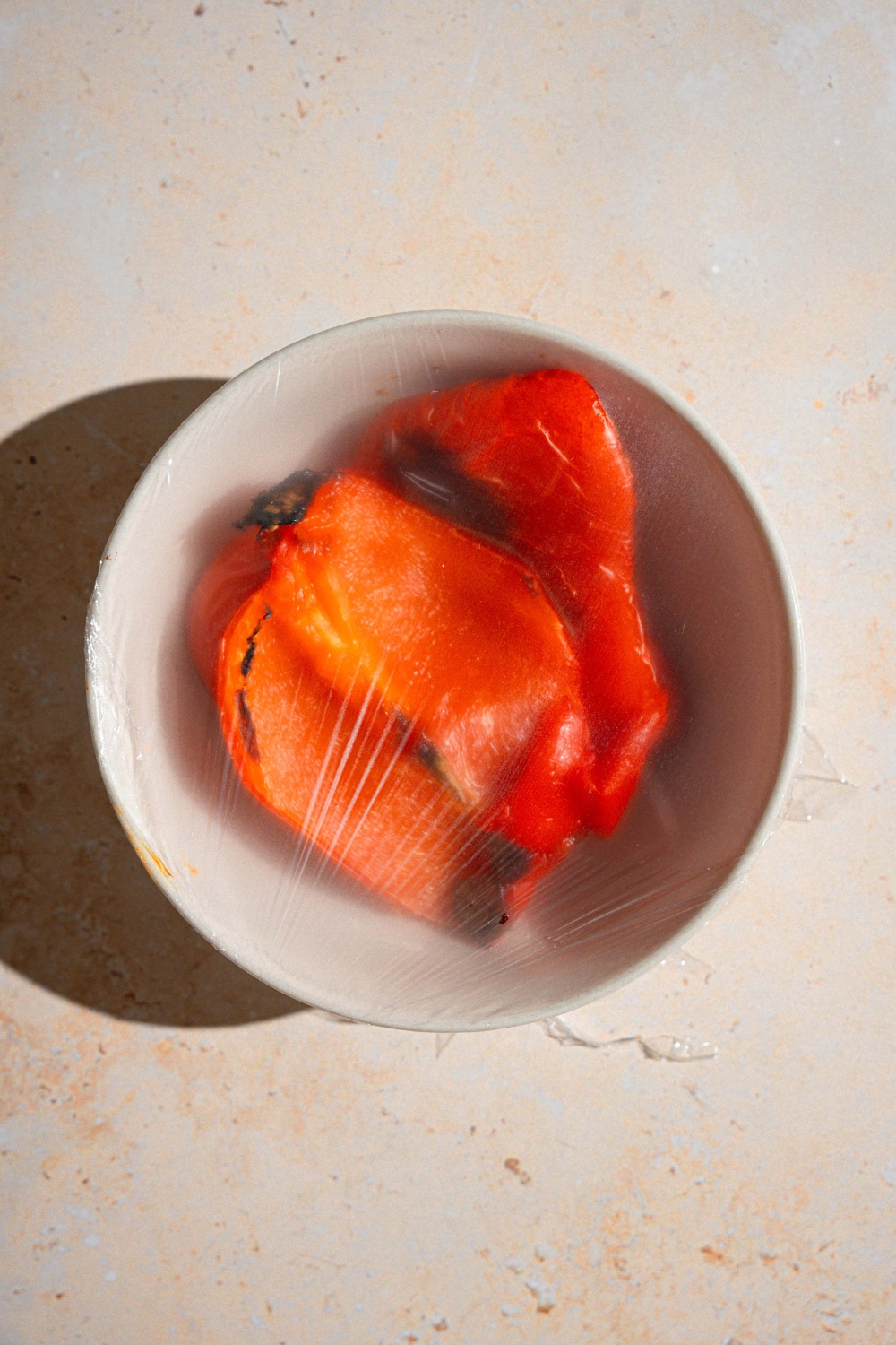 A bowl of steaming roasted red pepper wrapped in plastic wrap. The bowl is on a tan counter.