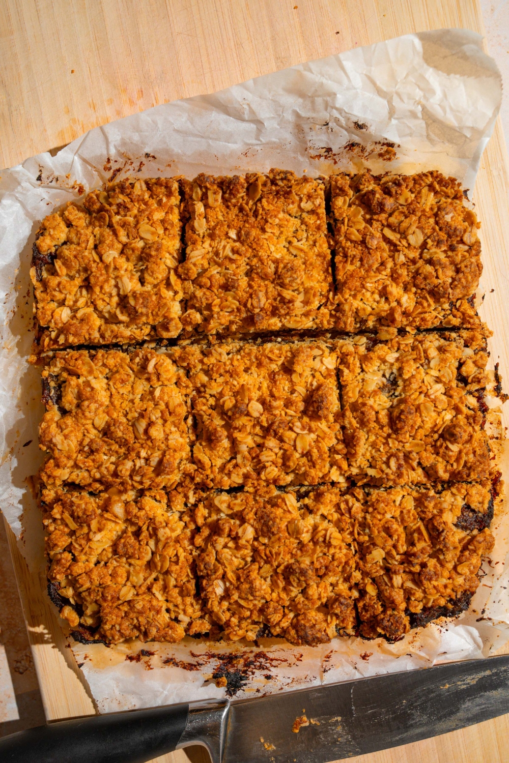 A wooden board with baked old fashioned date bars sliced in squares on a piece of parchment paper. There is a knife on the board.