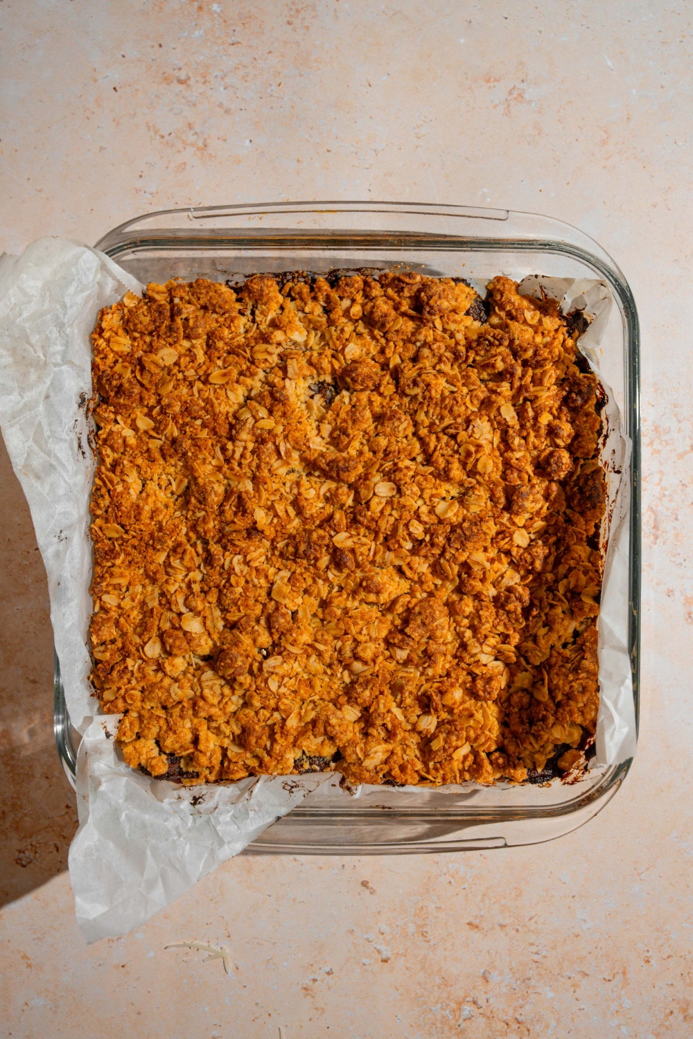 A glass baking dish lined with parchment paper with baked old fashioned date bars. The baking dish is on a tan counter.