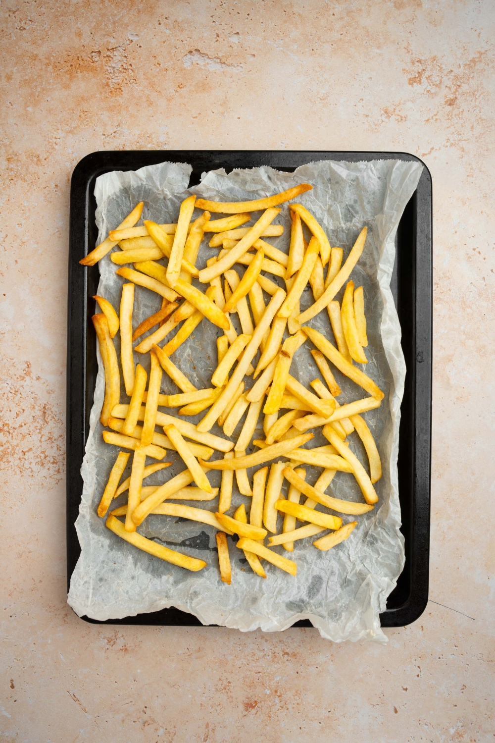 A baking sheet lined with parchment paper with baked french fries. The sheet is on a tan counter.