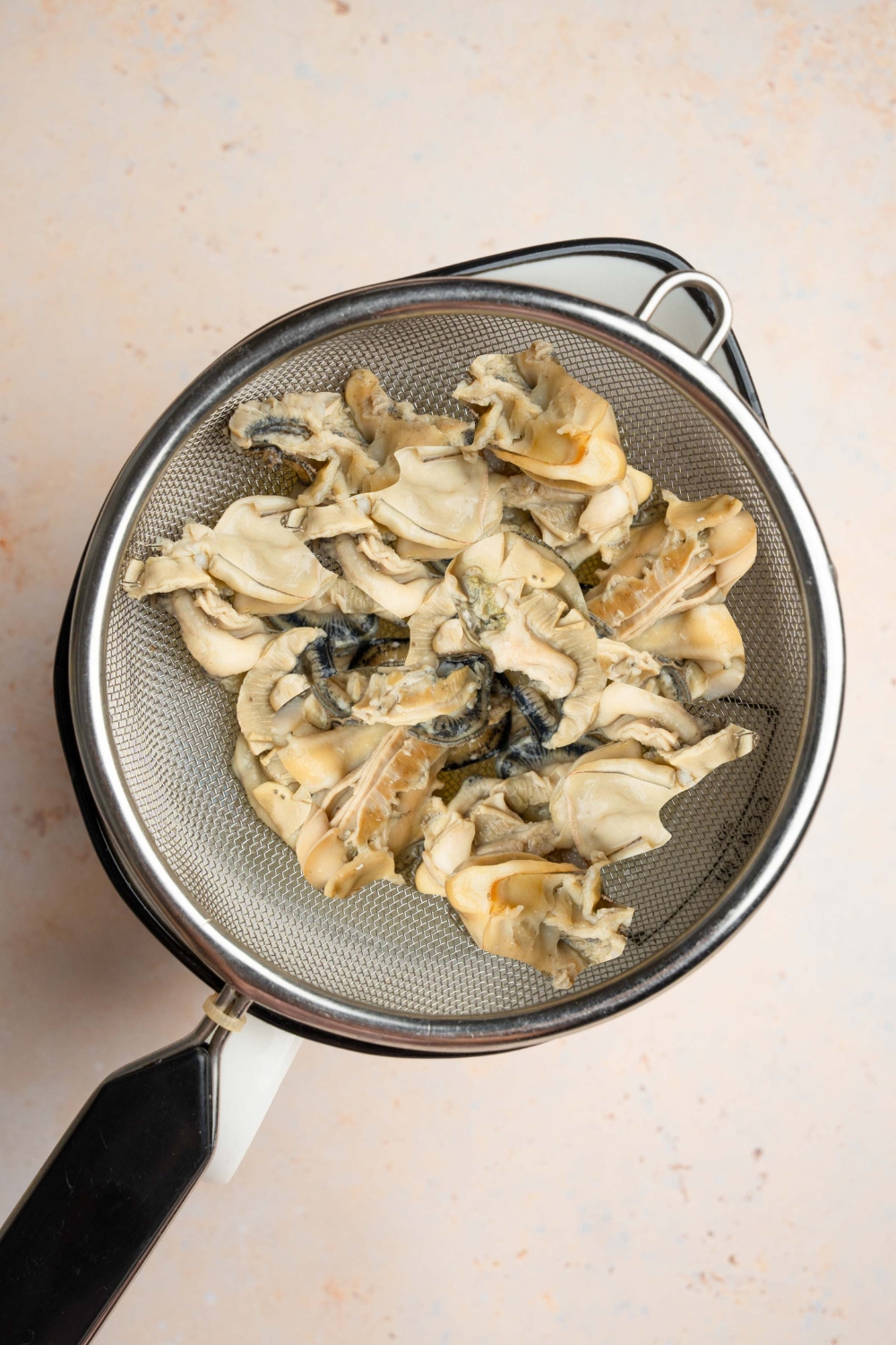 A sieve with strained oysters over a stock pot. The pot is on a tan counter.
