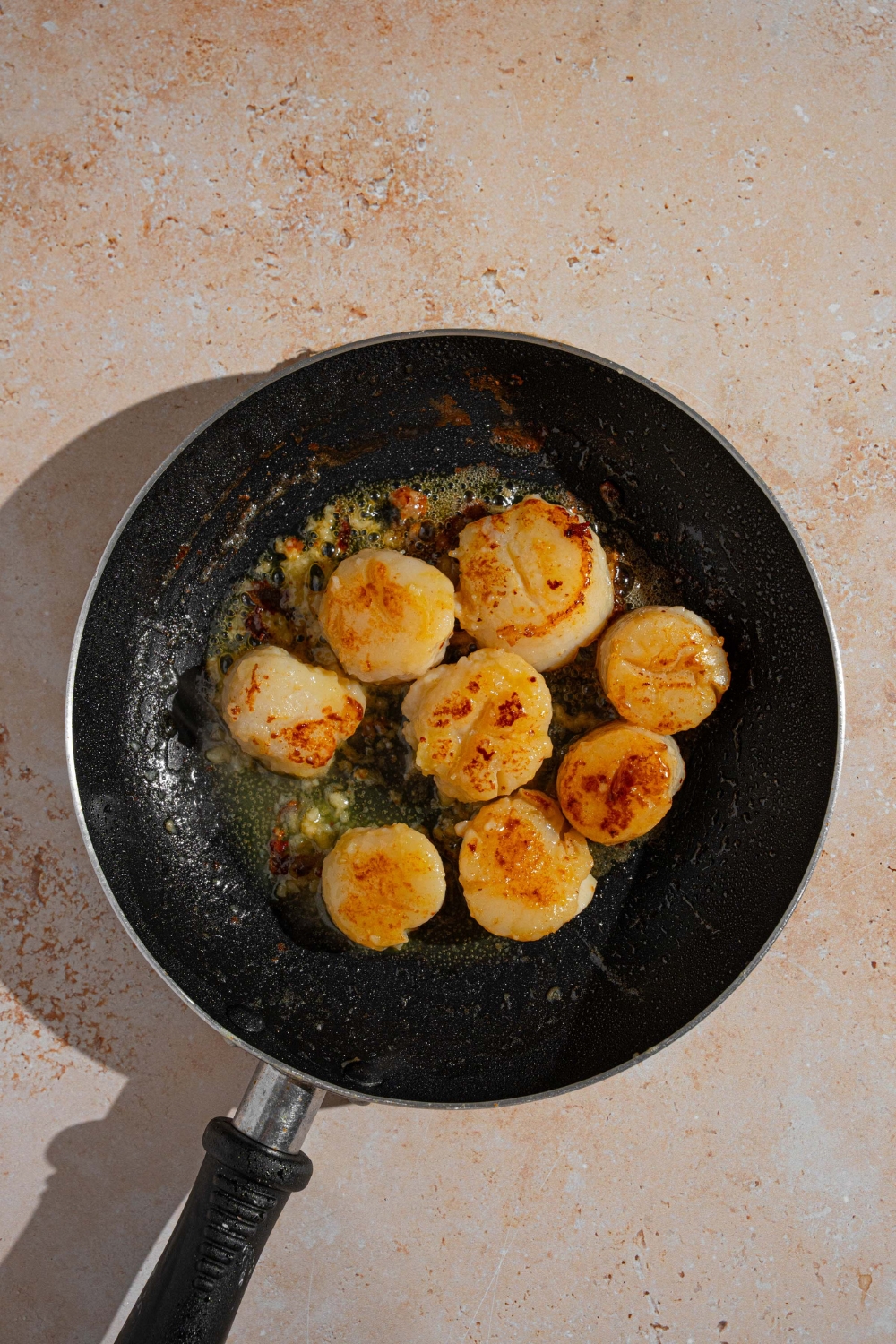 A skillet with scallops searing in butter and garlic. The skillet is on a tan counter.