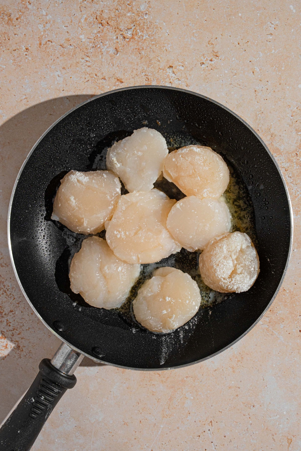 A skillet with seasoned scallops searing in butter and garlic. The skillet is on a tan counter.