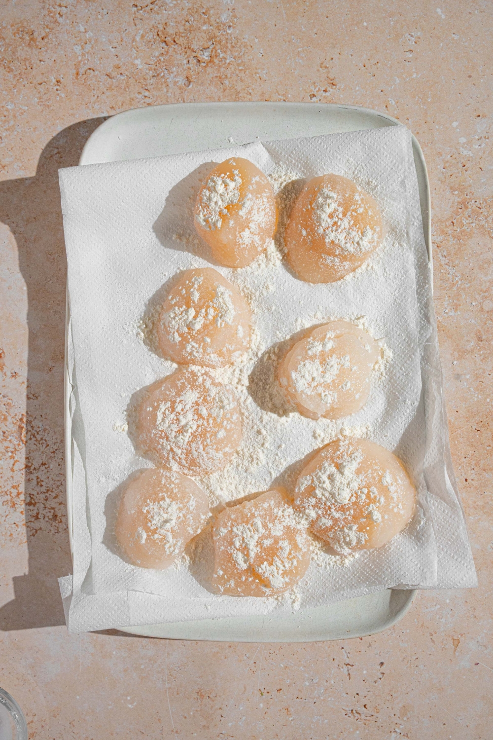 A white dish lined with parchment paper with defrosted scallops dusted with flour. The dish is on a tan counter.