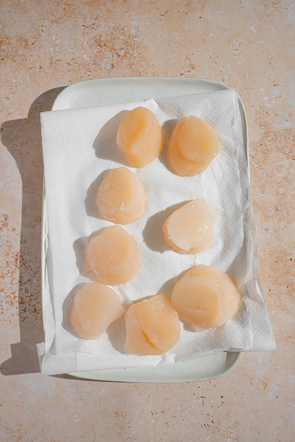 A white dish lined with parchment paper with defrosted scallops. The dish is on a tan counter.